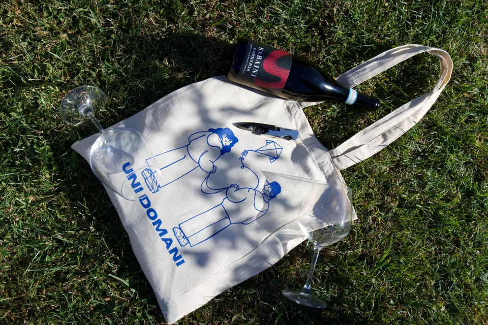 La borsa di tela di UniDomani sull’erba, con una bottiglia di Valpolicella, un cavatappi e due calici.